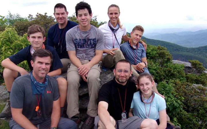 A group of parents and children pose for a photo on an overlook. There is a mountainous landscape in the background.